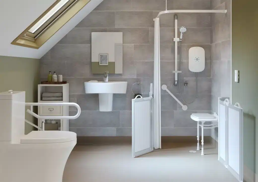 Accessible age care bathroom with a walk-in shower featuring a fold-down seat, grab bars, wall-mounted sink, and toilet with support rails. The sloped ceiling, skylight, and light gray tiled walls enhance the comfort of this wet room.