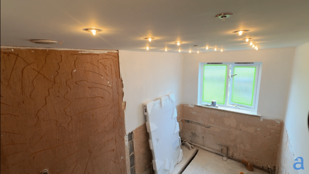 A bathroom under renovation with exposed plaster and brickwork on the walls. The ceiling features several newly installed recessed spotlights, and a white window with green protective film on the glass is visible at the far end of the room.