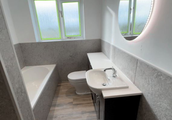 A modern finished bathroom with light grey marble-effect wall panels and wood-style flooring. The room features a white low level bathtub, a back-to-wall toilet, and a vanity unit with an integrated sink and chrome tap, situated beneath a large circular LED-illuminated mirror.