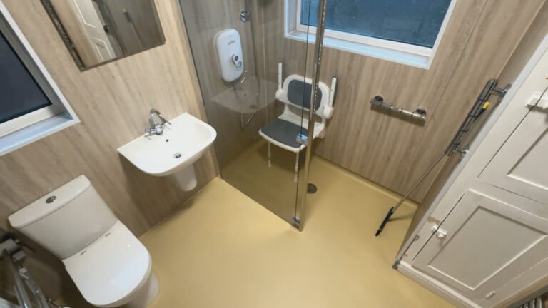 The finished level-access wet room, with a padded folding shower seat, glass screen, wall-hung basin and comfort-height toilet.