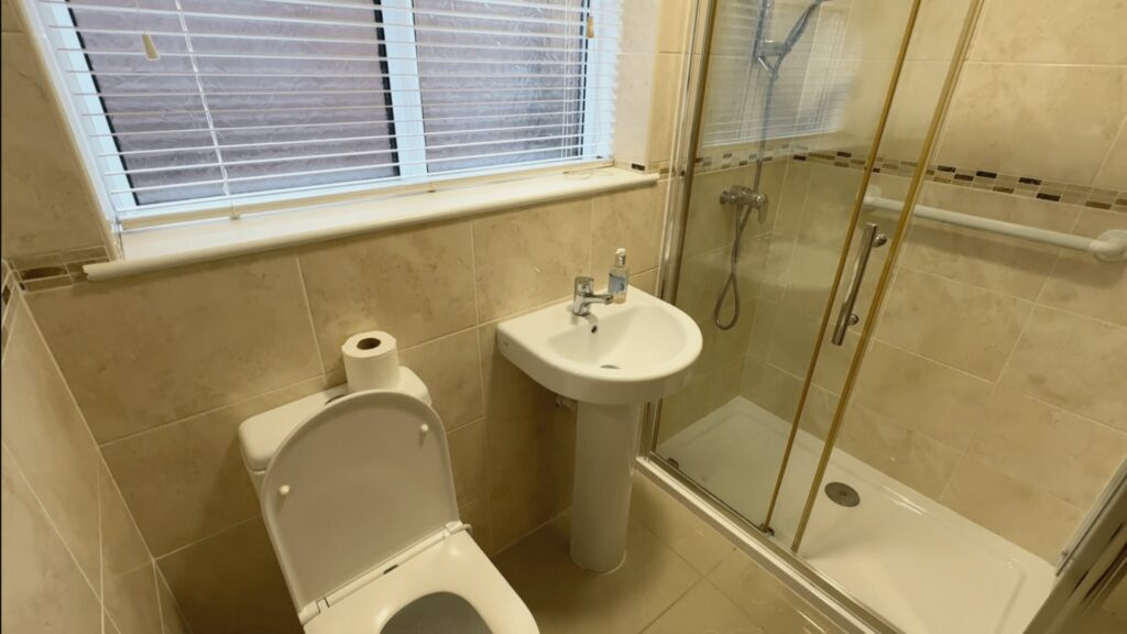 Steve's old bathroom before the work, with a raised shower tray, sliding glass doors, pedestal basin and beige tiling.