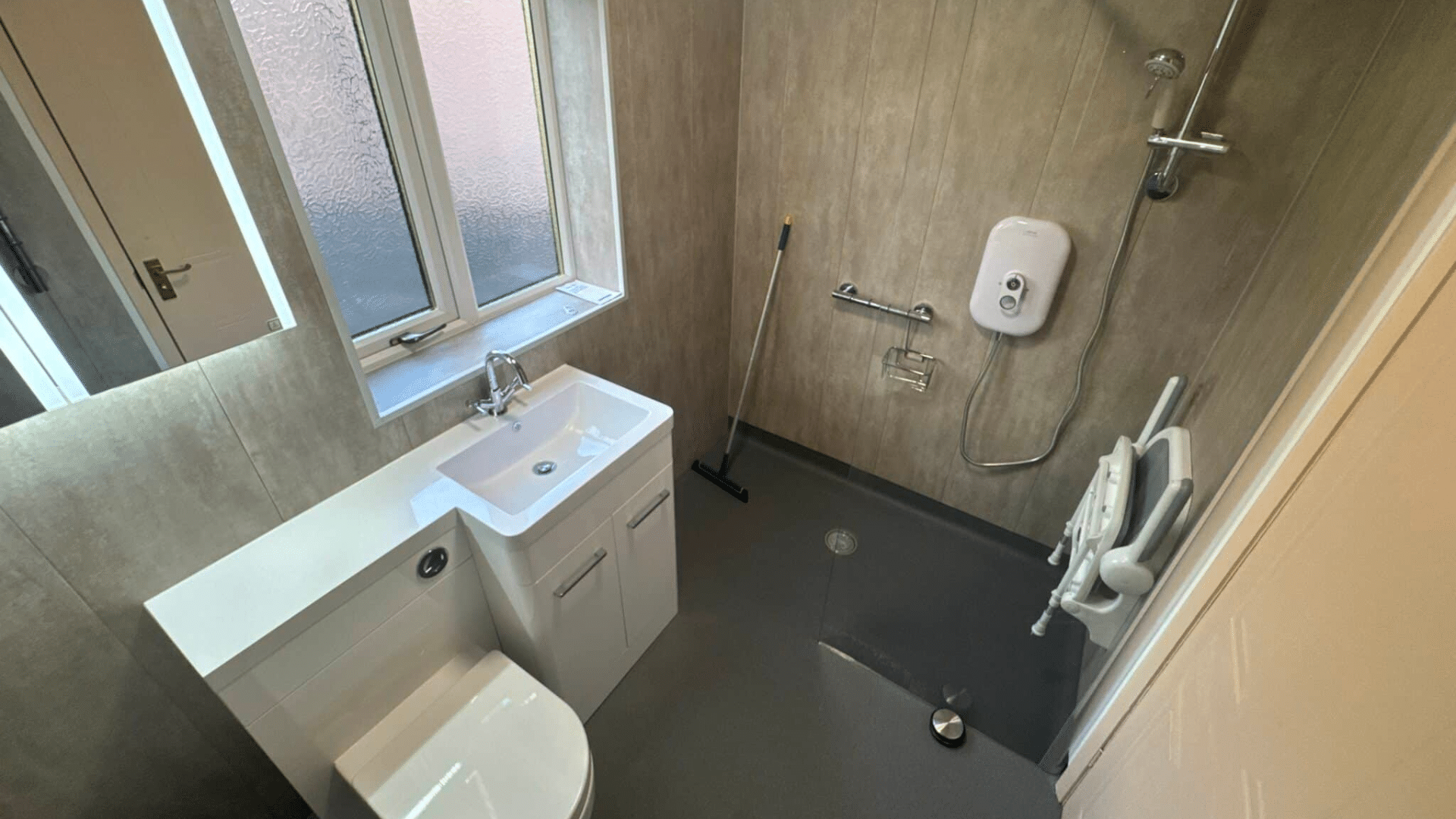 Accessible bathroom with a toilet, sink, and roll-in shower in a stylish wet room layout. The shower area features a wall-mounted shower seat, grab bars, and a handheld showerhead. A mop leans against the wall near a window.