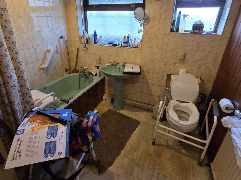 Bruce's old bathroom before the work, with a green bath, bath board, raised toilet seat, walking frame and carpeted floor.