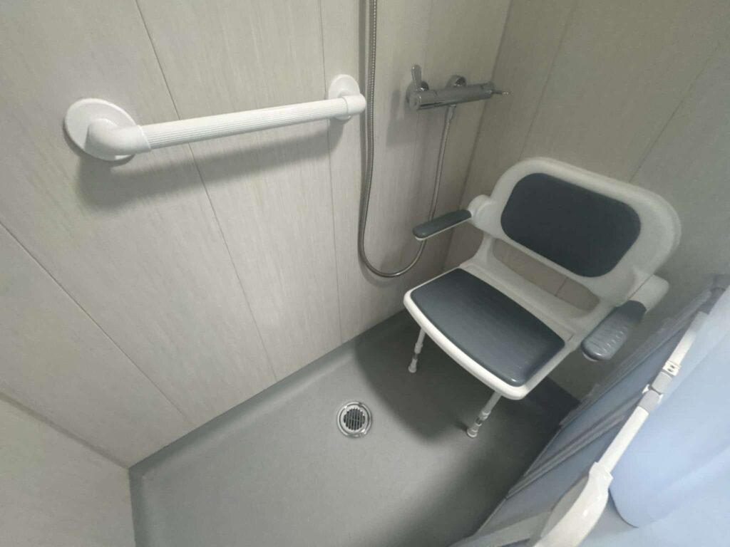 Close-up of the wall-mounted padded shower seat with armrests and horizontal grab rail in Room 1