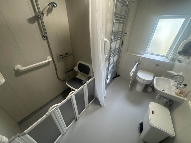 The finished wet room in Room 1, with a level-access shower, padded seat, white grab rails and grey non-slip flooring.