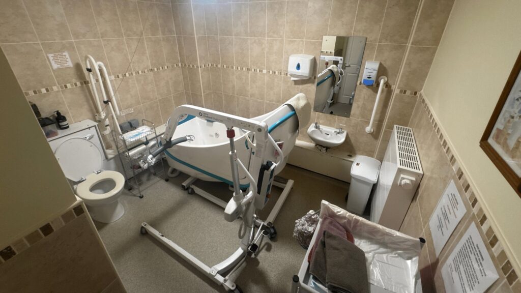 Room 2 at Horsfall House before the work, with a high-sided bath, mobile hoist and dated beige tiling