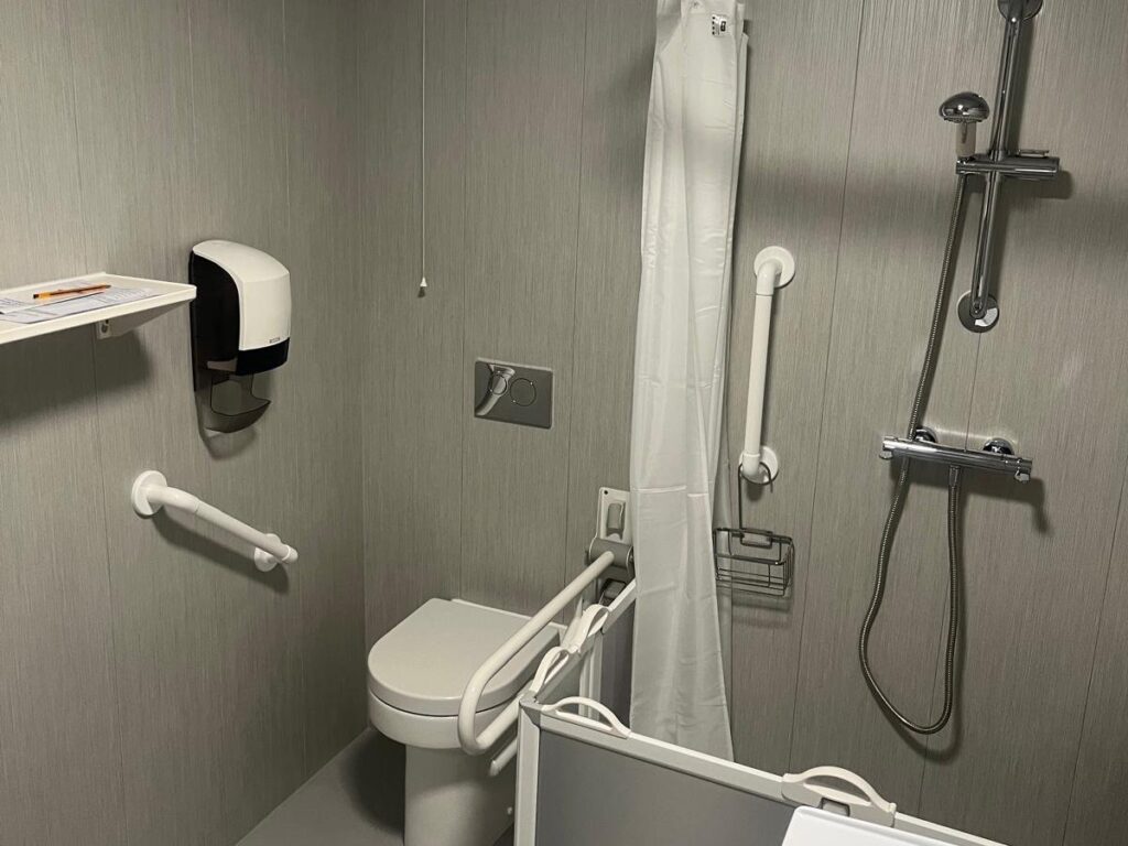 The toilet with a folding safety armrest in Room 2, beside the shower's half-height frosted carer doors.