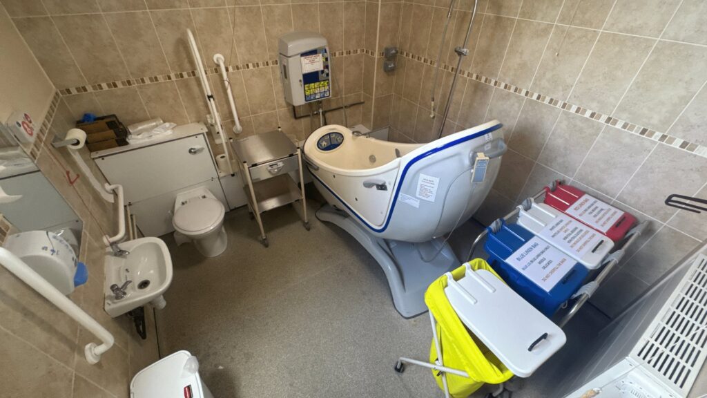 Room 3 at Horsfall House before the work, with dated beige tiling, an ageing assisted bath and laundry trolleys.