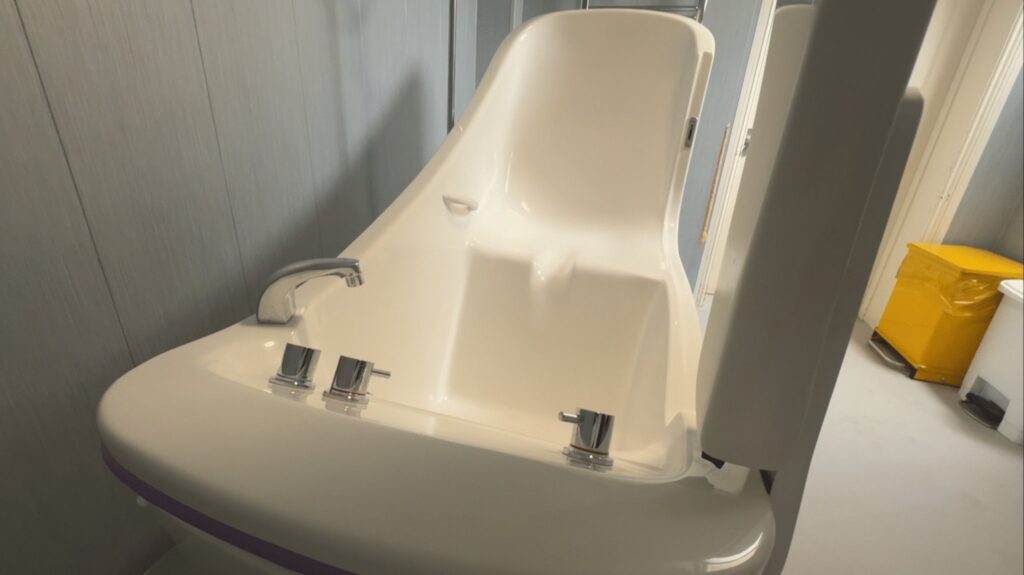 The toilet with its fold-down support rail in Room 3, against full-height easy-clean wall panels