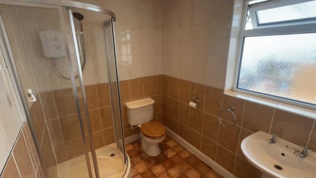 Lesley's old bathroom before the work, with a corner shower cubicle, step-up tray and dated brown tiling.
