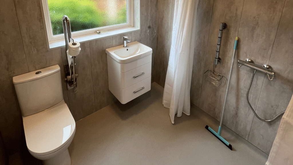 A wide-angle view of a completed accessible wet room featuring natural stone-effect waterproof wall panels and slip-resistant light grey safety flooring. The layout includes a white close-coupled toilet on the left, a small wall-hung vanity unit with a white washbasin in the centre, and a level-access walk-in shower area on the right. A vertical chrome safety grab rail with a toilet roll holder is mounted next to the toilet, and a second vertical grab rail with a soap dish is positioned within the shower area. A white shower curtain is partially drawn, and a window provides a view of green foliage outside.