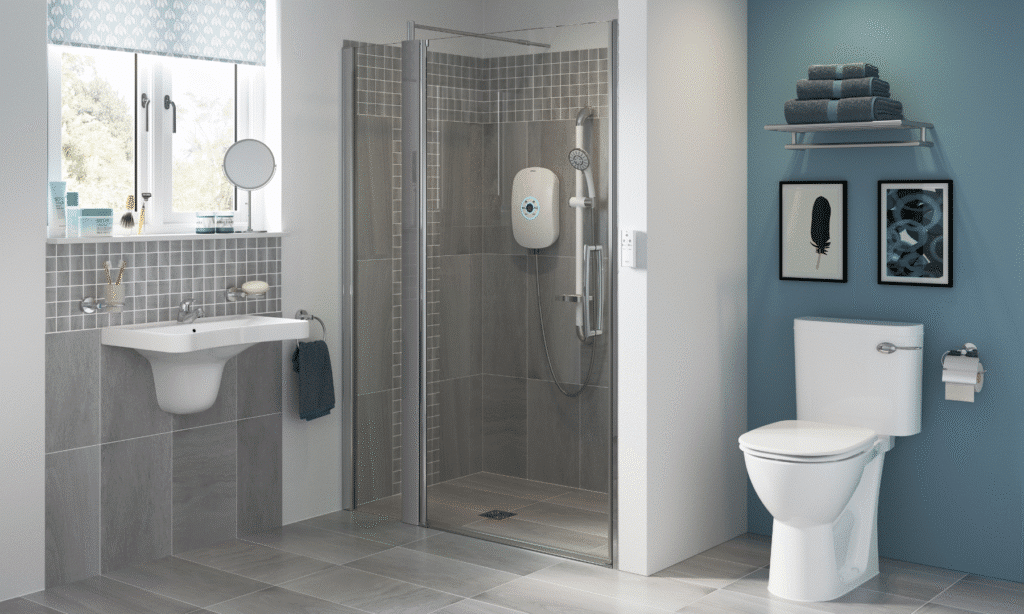 A bright, modern mobility bathroom featuring a combination of blue and white walls with large grey floor tiles. The layout includes a level-access shower with grey wall tiles, a white electric shower unit, and an integrated chrome safety grab rail. To the left, a white semi-pedestal washbasin is mounted against a grey tiled splashback, and to the right, a white close-coupled toilet is positioned against a blue feature wall. A large window provides natural light, and a chrome towel rack with blue towels is mounted above the toilet.