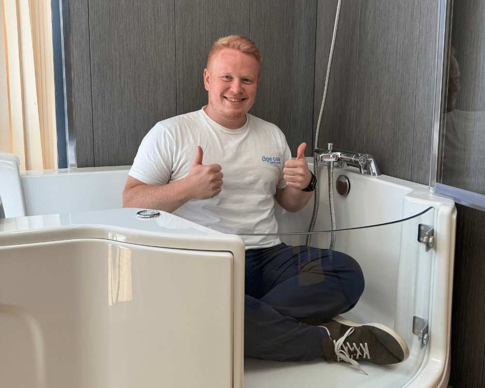 A smiling man with short blond hair sits cross-legged in a modern walk in bath, giving two thumbs up. He is wearing a white t-shirt, dark pants, and sneakers. The age care bathrooms feature a glass door and sleek fixtures for safety and comfort.
