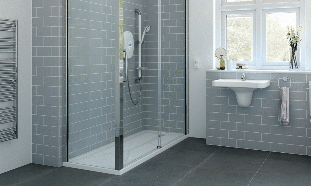 Modern bathroom with grey tiled walls, a walk-in glass shower perfect for Age Care Bathrooms, wall-mounted electric shower, white sink, towel rail, mirror, and window letting in natural light. Minimalist and clean design.
