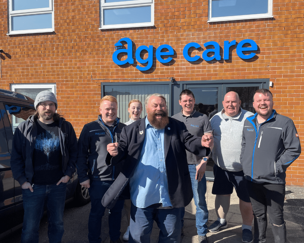 A group of seven people, smiling and standing outside a brick building with a blue age care sign above the entrance, enjoying a sunny day together—where features like walk in showers make comfort and safety a priority.
