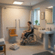 A person in a wheelchair smiles as they are about to use a level-access shower in a modern, accessible wet room in a UK home. The room is comfortable and well-lit, with grab rails, a fold-down shower seat, non-slip flooring, and towels within easy reach.