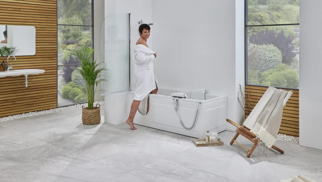 A woman in a white bathrobe stands on a stone tile floor, stepping into a white walk-in bathtub. The modern bathroom has a wood-paneled wall, a large window looking out onto a garden, and a small sink.