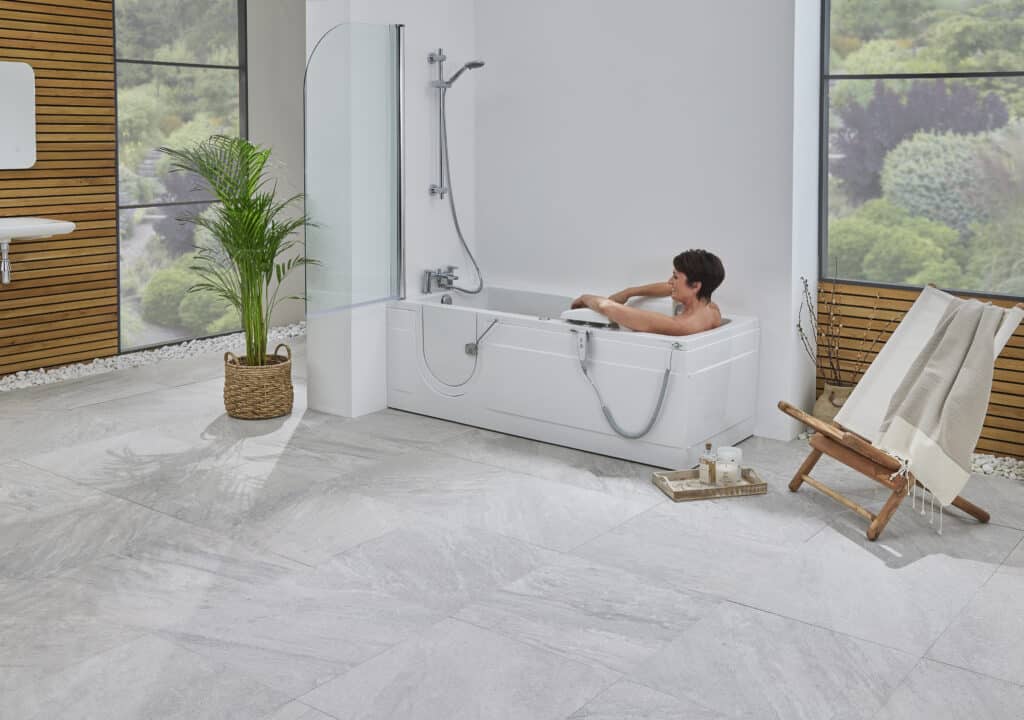 A person relaxes in a modern white walk-in bathtub with a clear glass shower screen, set in a bright bathroom with light grey floor tiles and a wood-paneled wall.