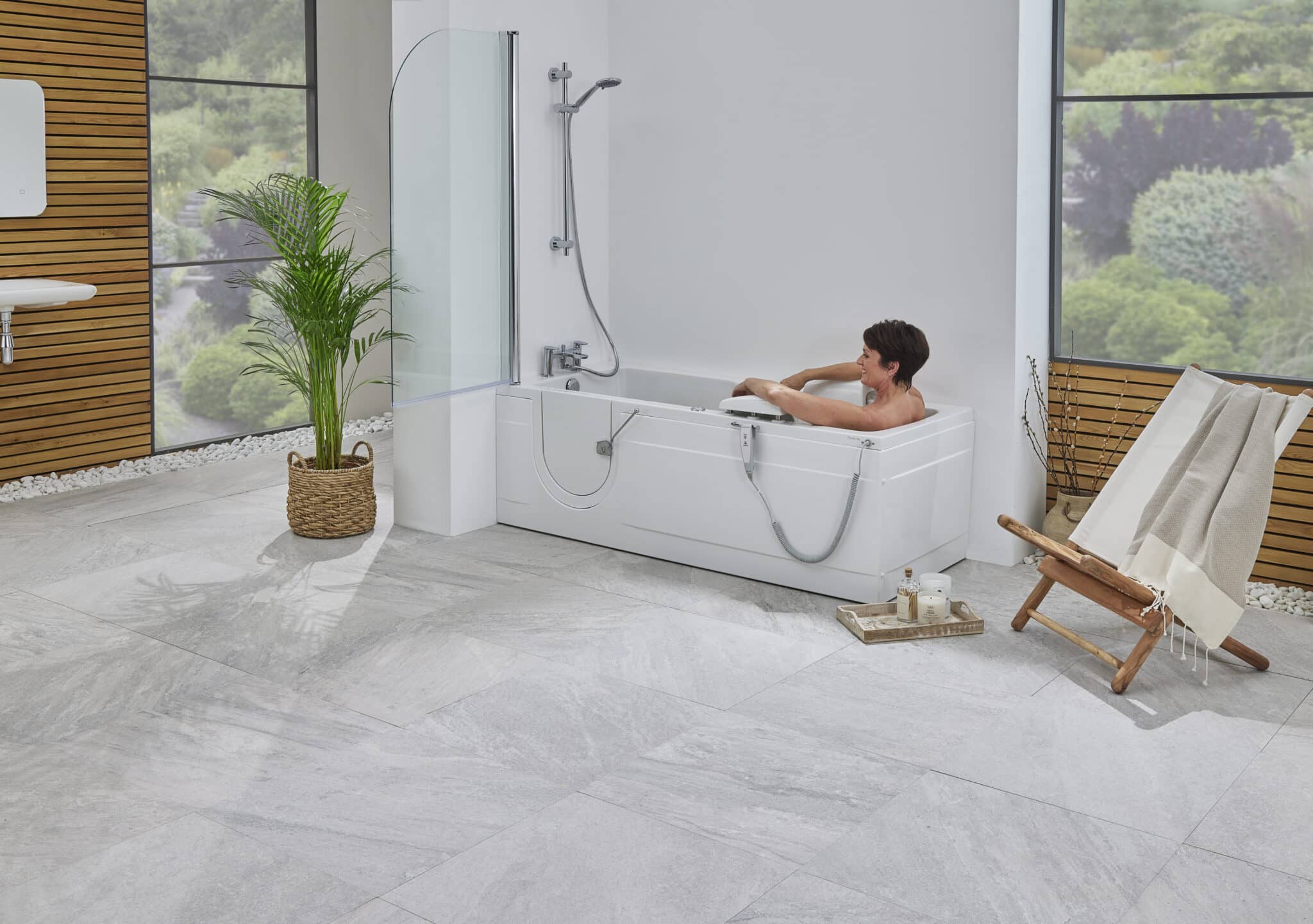A person relaxes in a modern white walk-in bathtub with a clear glass shower screen, set in a bright bathroom with light grey floor tiles and a wood-paneled wall.