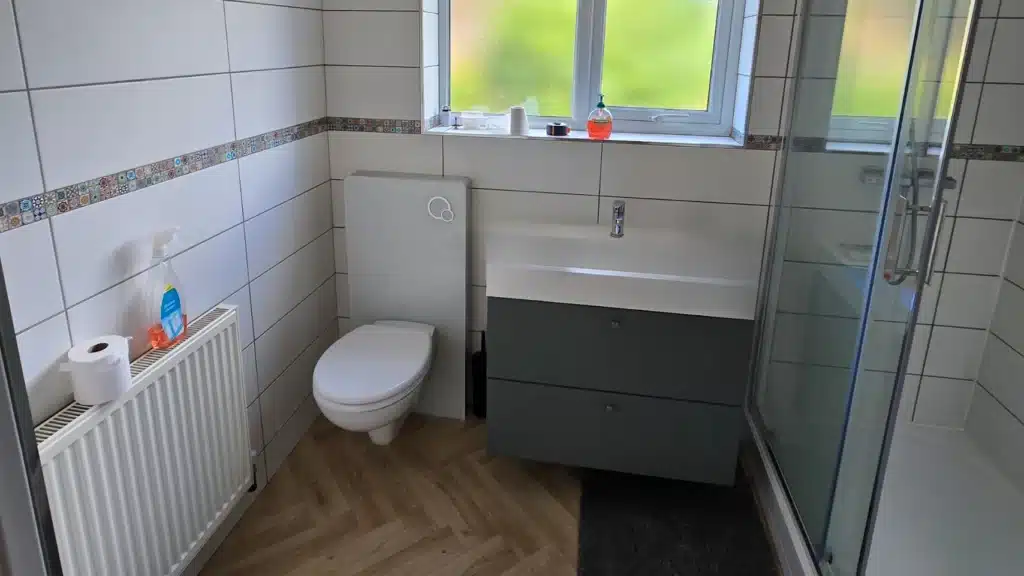 A bathroom before renovation, featuring white tiled walls with a decorative grey mosaic border and wood-effect herringbone flooring. The room includes a back-to-wall toilet, a modern grey vanity unit with a large white washbasin, and a standard glass shower cubicle with a high-profile white tray. A white radiator is positioned against the left wall, and a large window provides a view of green foliage outside.
