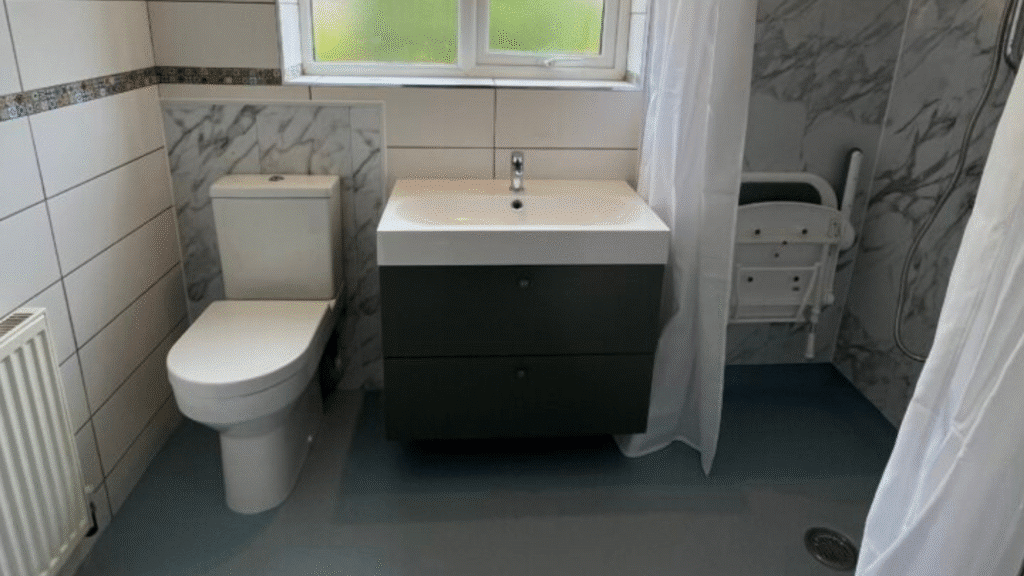 Sandra’s New Accessible Bathroom features a modern design with a toilet, a sink with a dark cabinet, a folding shower seat, slip-resistant flooring, tiled walls, and a window above the sink for natural light.