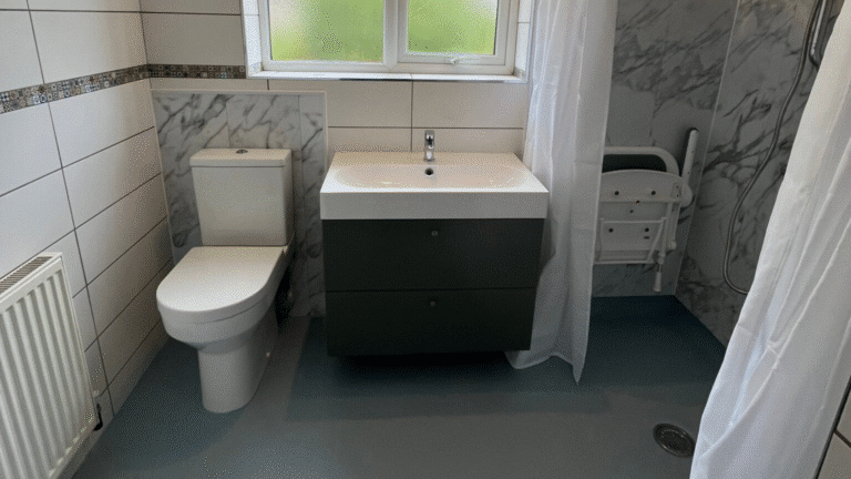 A completed accessible wet room renovation featuring grey marble-effect waterproof wall panels in the shower area and slip-resistant blue safety flooring. The layout includes a white close-coupled toilet, a modern grey vanity unit with an integrated washbasin, and a level-access walk-in shower with a wall-mounted folding seat and a white shower curtain. Original white wall tiles with a mosaic border are preserved on the left, and a frosted window provides natural light.