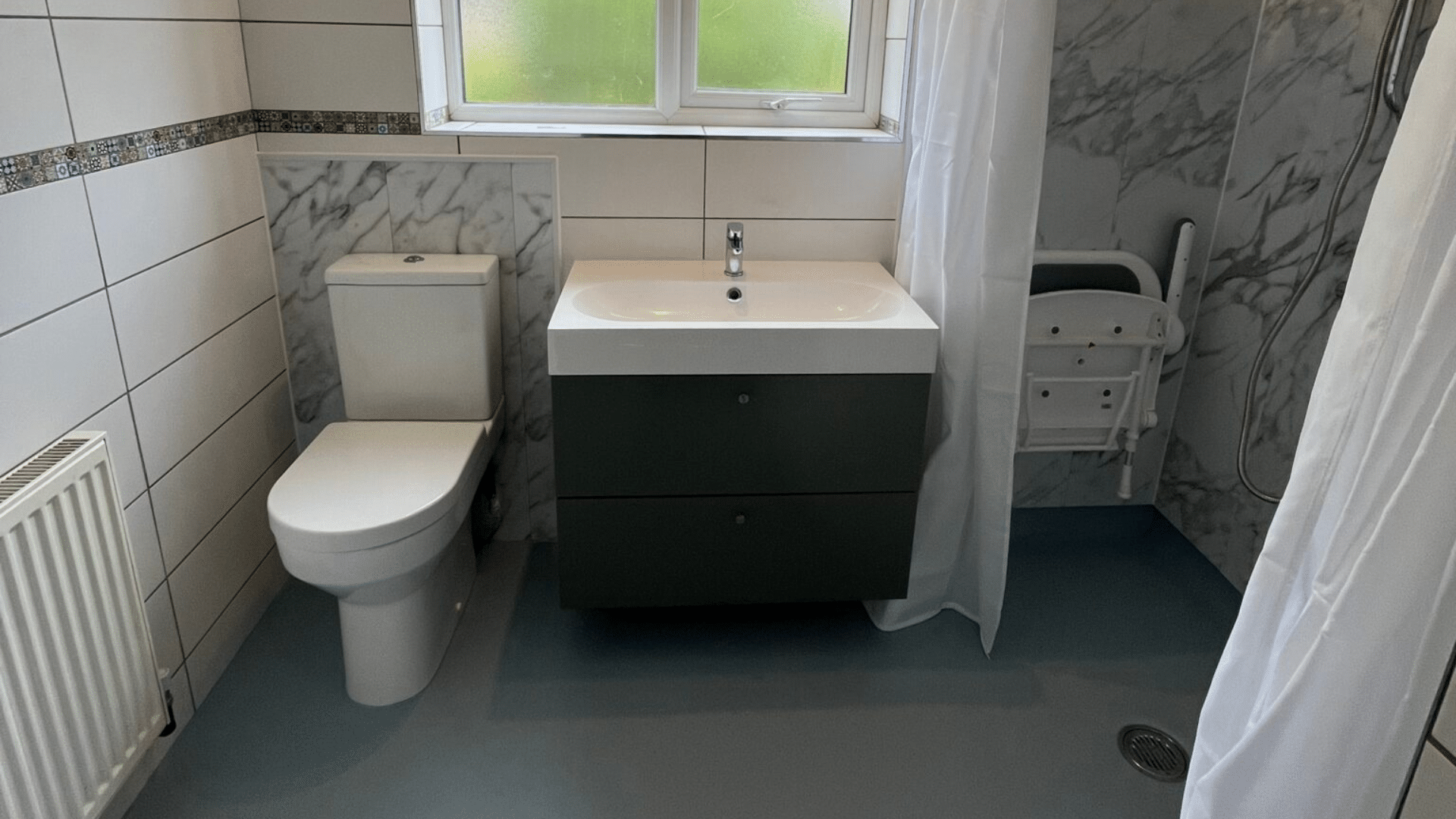 A completed accessible wet room renovation featuring grey marble-effect waterproof wall panels in the shower area and slip-resistant blue safety flooring. The layout includes a white close-coupled toilet, a modern grey vanity unit with an integrated washbasin, and a level-access walk-in shower with a wall-mounted folding seat and a white shower curtain. Original white wall tiles with a mosaic border are preserved on the left, and a frosted window provides natural light.