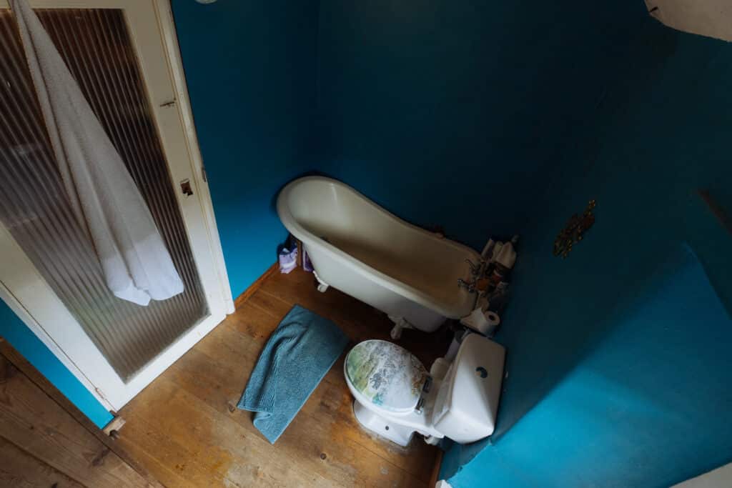A top-down, wide-angle view of a traditional bathroom before renovation, featuring a cream-coloured roll-top bathtub and a white toilet. The room has deep teal-painted walls, light wood-effect flooring, and a glass-panelled wooden door partially covered by a white towel. A blue towel lies on the floor next to the toilet.
