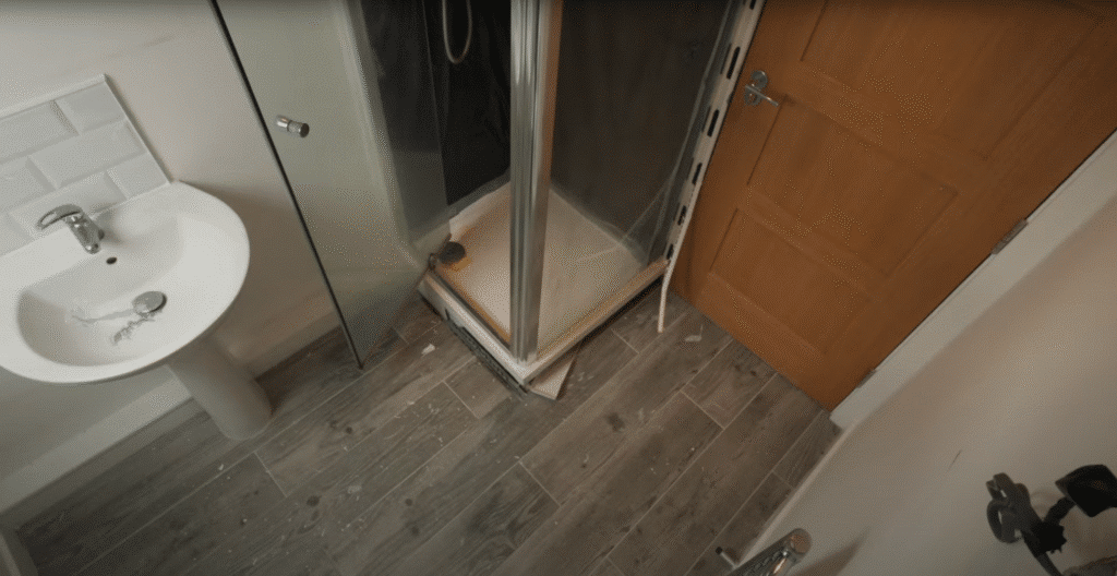 A top-down, bird's-eye view of a compact bathroom before renovation, featuring a white pedestal washbasin with a chrome mixer tap and a standard corner shower cubicle with a high-profile white tray. The room has grey wood-effect floor tiles and a light wood-finish door. A white safety grab rail is visible in the bottom-right corner, mounted next to the doorway.