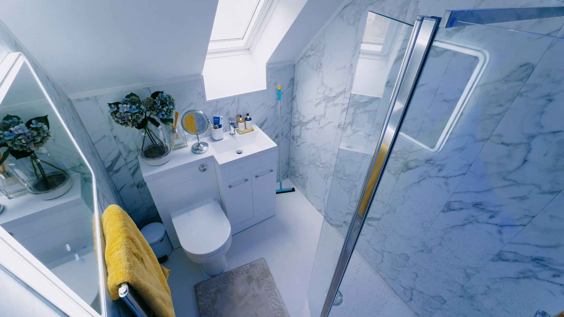 A compact upstairs wet room installation by Age Care Bathrooms featuring marble-effect wall panels, a level-access glass shower screen, and space-saving fixtures under a sloped loft ceiling with a skylight.