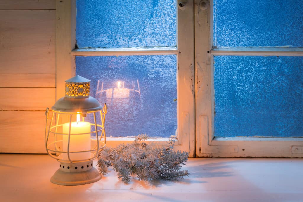 Warm candle lantern glowing beside a frosted window on a winter evening.