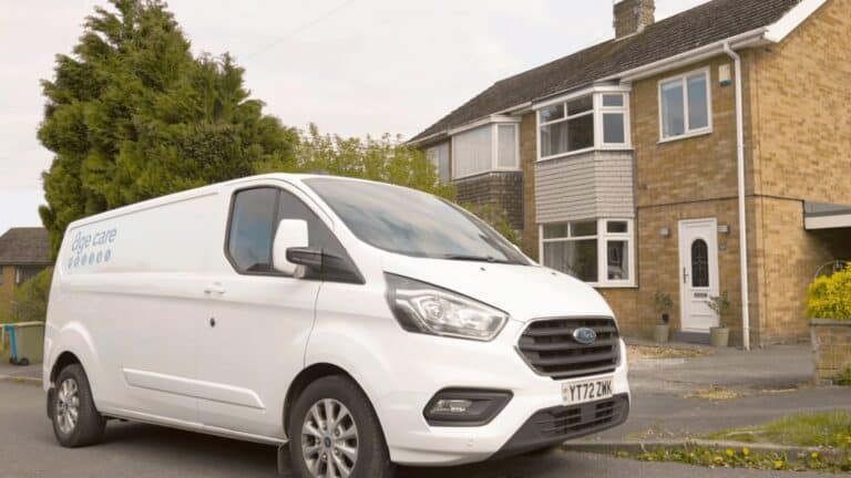 A white van with eye care branding is parked on the street in front of a two-story brick house, featuring a sloped roof and a small front yard—ideal for homeowners interested in Buy Now Pay Later Bathrooms.