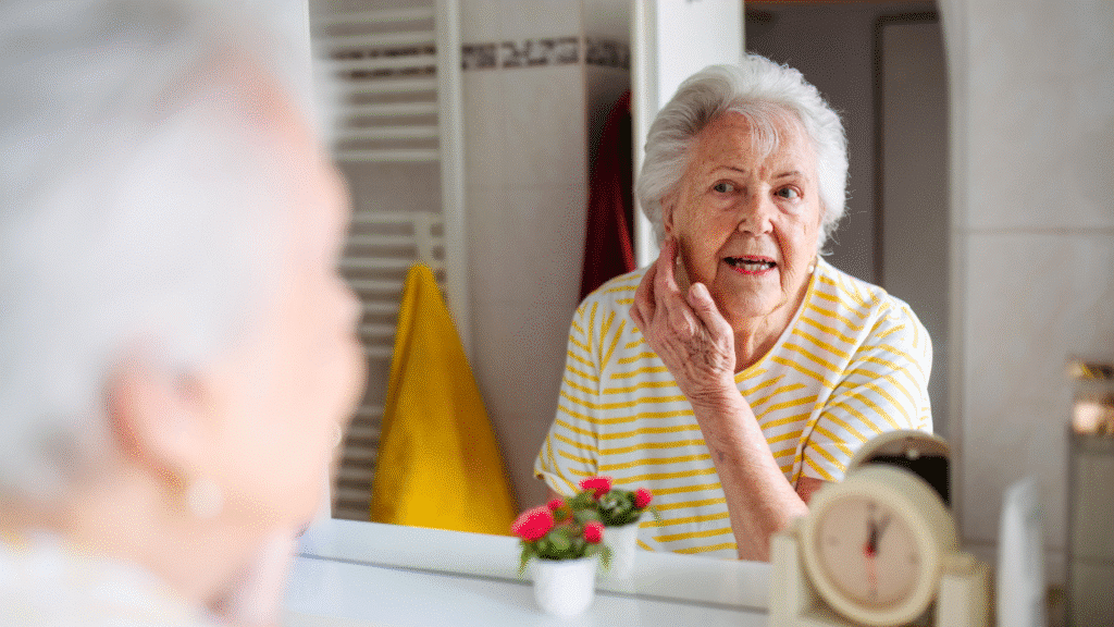 An elderly woman with gray hair, wearing a yellow striped shirt, looks at herself in the bathroom mirror while touching her cheek. A small plant and a clock are visible on the countertop in this Age Care Bathrooms setting.