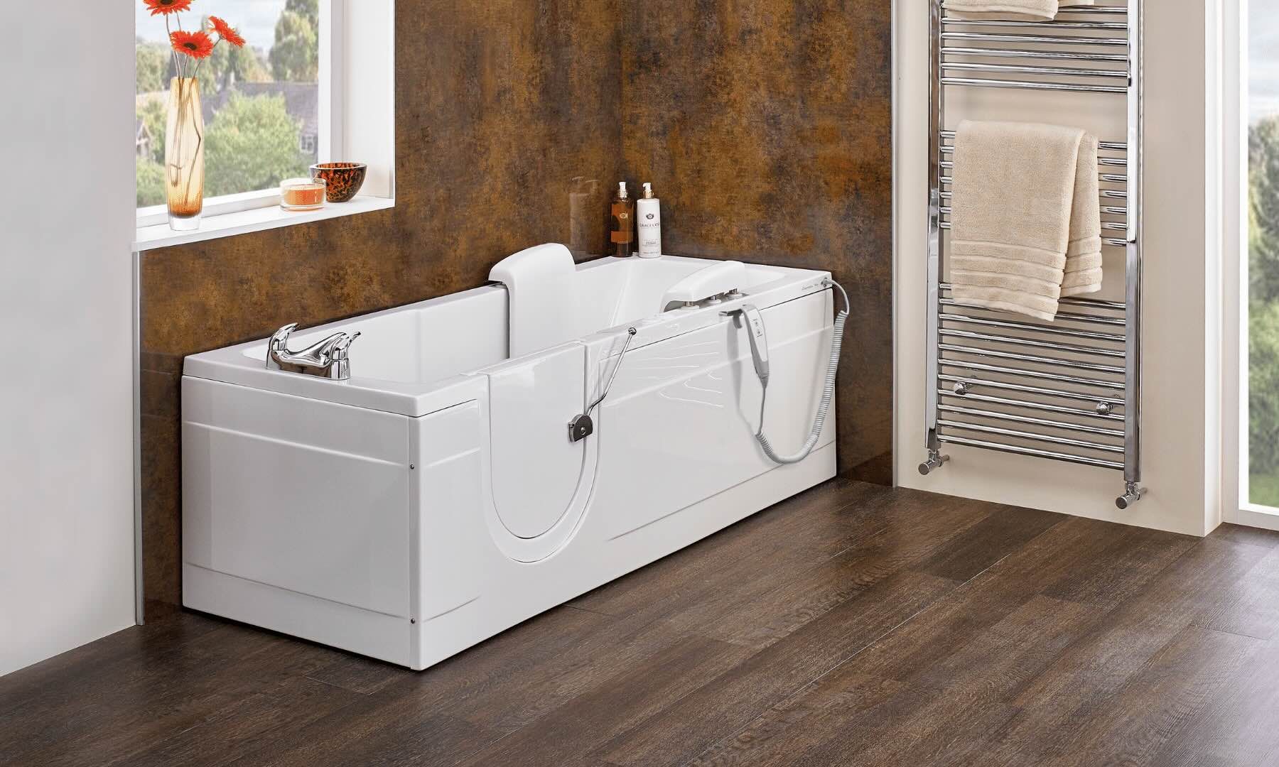 A modern bathroom with a white walk-in bathtub, grab bars, showerhead, and a pillow; brown textured walls, dark wood flooring, a window with flowers, and a chrome towel rack with folded towels.