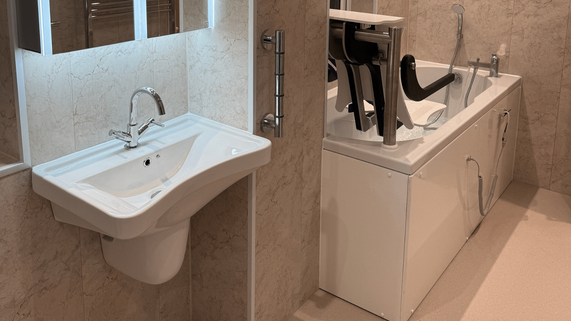 A modern, completed accessible bathroom featuring neutral-toned wall tiles and slip-resistant flooring. The layout includes a wall-hung washbasin with a chrome mixer tap and an illuminated mirror cabinet above it. To the right, a white bathtub is fitted with an integrated powered seat lift to assist with entry and exit. A chrome vertical grab rail is mounted to the wall between the basin and the bath for additional support.