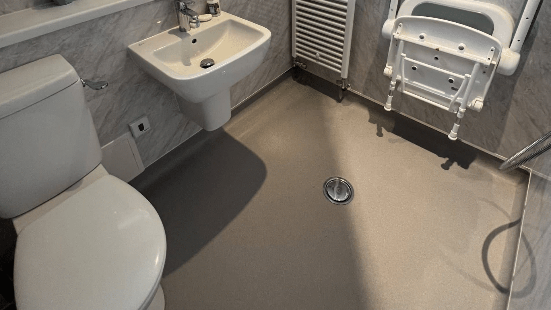 A top-down view of a completed mobility bathroom renovation, showcasing a light brown, slip-resistant safety floor that is completely level and flush with a central chrome drainage grate. The flooring has an integrated coved skirting that runs up the base of the marble-effect wall panels to create a watertight seal. The layout also includes a white close-coupled toilet, a wall-hung washbasin, and a wall-mounted folding shower seat with grey padding.