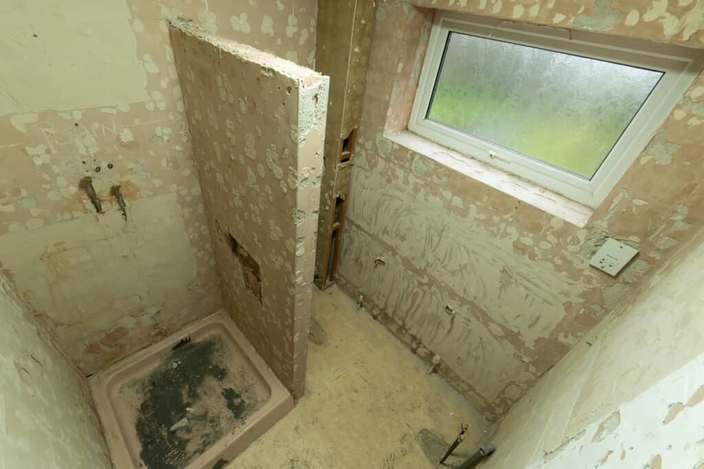 Worn bathroom prior to renovation, showing a small shower area with a low tray and partial partition, exposed and damaged walls with stripped plaster, and a window above. The space appears dated and in need of a full accessible refurbishment.