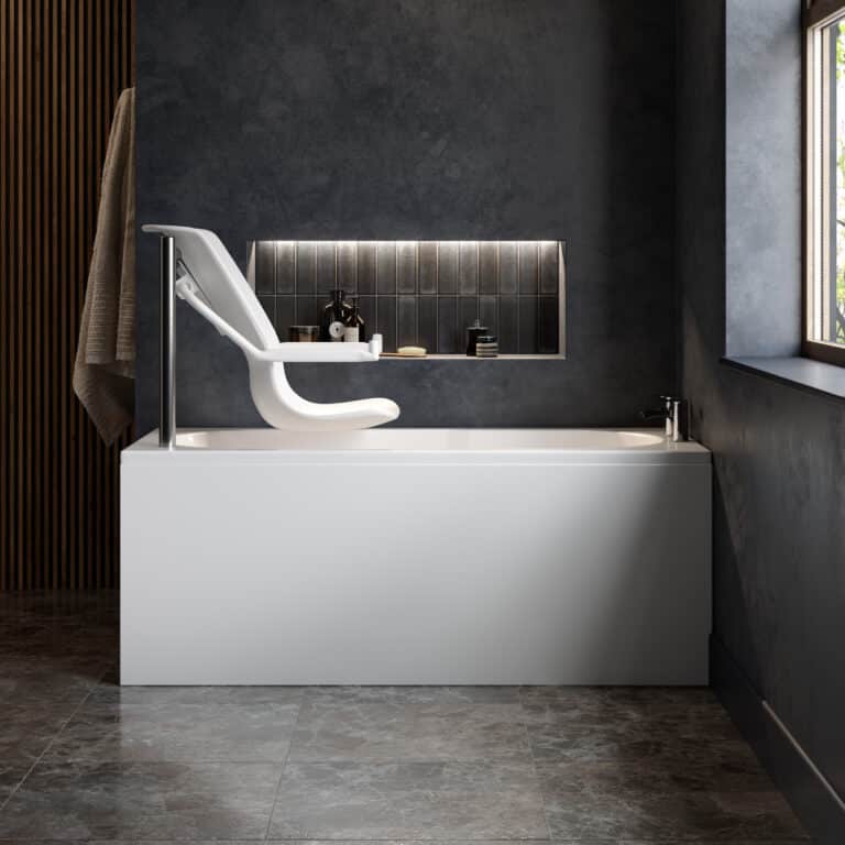 A side-angle view of a modern accessible bathroom featuring an Omega walk-in bath with the powered seat lift in its raised, entry position. The white contoured seat is elevated and tilted forward over the edge of the bath, supported by a vertical chrome pillar. The setting includes dark grey textured walls, marble-effect floor tiles, and a lit recessed shelf containing toiletries.
