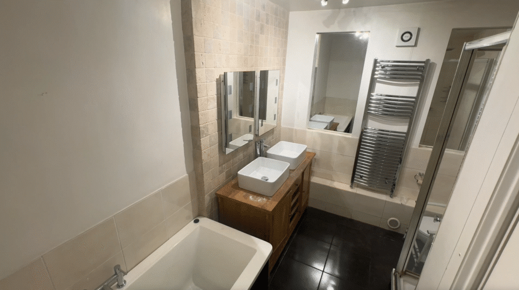 Before photo of a standard bathroom featuring a white bathtub, a wooden vanity with double square vessel sinks, and a tall chrome heated towel rail, prior to an Age Care Bathrooms mobility transformation.
