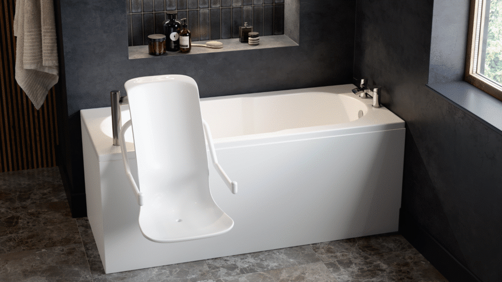 A modern accessible bathroom featuring an Omega walk-in bath with a powered seat lift. The white rectangular bath is equipped with a high-backed, contoured powered seat and integrated armrests that extend over the side for easy entry. The room has dark grey textured walls with a recessed shelf for toiletries and large grey marble-effect floor tiles. A large window on the right provides natural light, and a neutral-toned towel hangs on the left wall.