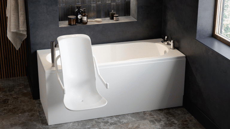 A modern accessible bathroom featuring an Omega walk-in bath with a powered seat lift. The white rectangular bath is equipped with a high-backed, contoured powered seat and integrated armrests that extend over the side for easy entry. The room has dark grey textured walls with a recessed shelf for toiletries and large grey marble-effect floor tiles. A large window on the right provides natural light, and a neutral-toned towel hangs on the left wall.