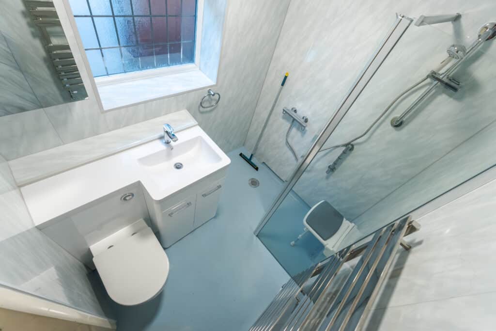 After photo of a modern accessible wet room by Age Care Bathrooms, featuring blue non-slip safety flooring, a white vanity unit with integrated toilet and sink, and a walk-in shower with a glass screen and fold-down seat.