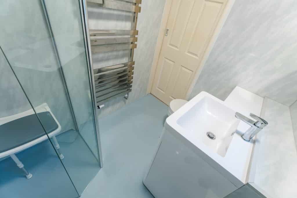 High-angle view of a completed accessible bathroom by Age Care Bathrooms, featuring a walk-in shower with a fold-down seat, blue non-slip safety flooring, a white vanity unit with a chrome lever tap, and a heated towel rail.