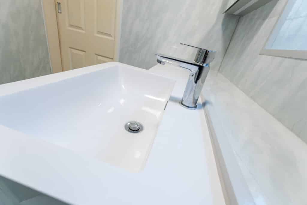Close-up angled view of a modern white ceramic vanity basin with a polished chrome lever-style mixer tap, showcasing easy-to-operate fixtures for an accessible Age Care Bathroom installation. Background shows grey tiled walls and a wood door.