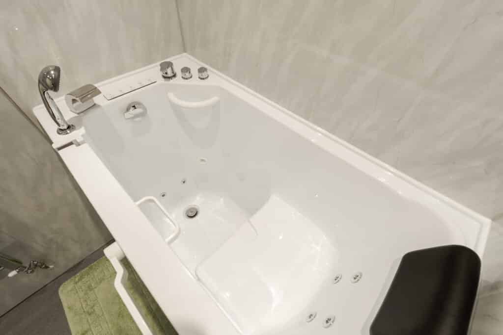 High-angle close-up of a luxury walk-in whirlpool bath installed by Age Care Bathrooms, featuring a black padded headrest, integrated spa jets, a hand-held shower attachment, and chrome controls.