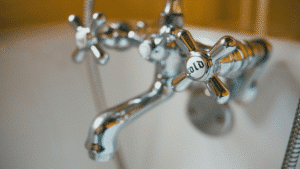 A close-up, selective-focus photograph of a highly polished, vintage-style chrome bathroom faucet assembly. The focus is tightly on a foreground 'cross-head' (four-spoke) handle, which features a central white ceramic disc clearly inscribed with the word 'COLD' in black. A main curving faucet spout and a flexible metal shower hose are in the blurred background against a white porcelain bathtub.