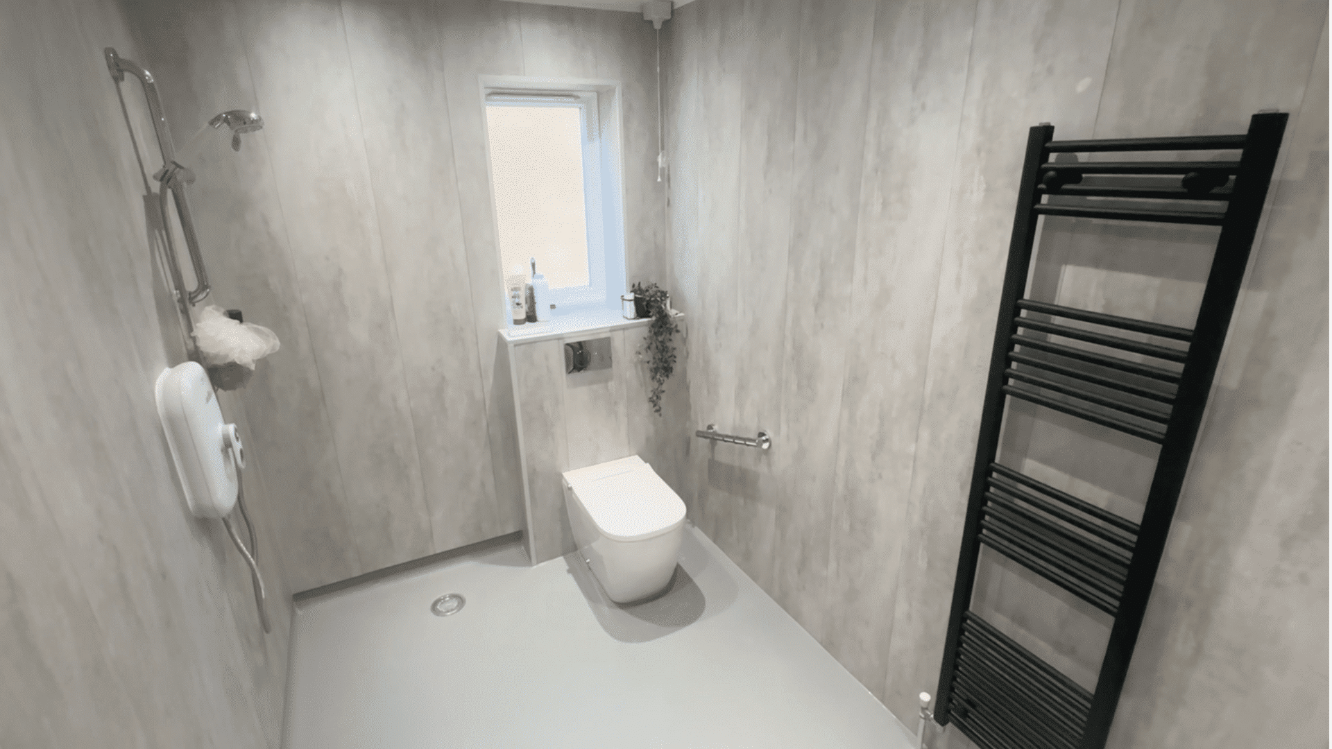 A high-angle photograph showcasing a completed, modern wet room installation by Age Care Bathrooms. Seamless grey non-slip flooring provides a barrier-free, level-access environment. Features include a walk-in shower on the left with non-slip flooring, a central toilet with a chrome grab rail, wood-effect wall paneling throughout, and a prominent black ladder towel radiator on the right wall, demonstrating a safe, stylish, and dignified home adaptation.
