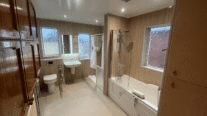 A detailed, wide-angle photograph of a newly installed, fully accessible bathroom designed for mobility and safety. On the right, a modern walk-in bathtub-and-shower combination is set against light wood-effect paneled walls and next to a large privacy-glass window. The tub features a transparent glass door, a built-in white transfer seat, and both overhead and handheld showerheads. On the left side, wooden storage cabinets are seen above a comfort-height raised toilet with an adjacent wall-mounted grab rail. Further down, there is a wall-mounted pedestal sink with a mirror above it, flanked by two more privacy-glass windows. In the far background, a curtained showering area and a partial enclosure are visible. The entire room has a matching light wood paneling on the walls and a light grey slip-resistant floor. The ceiling features recessed spotlights.