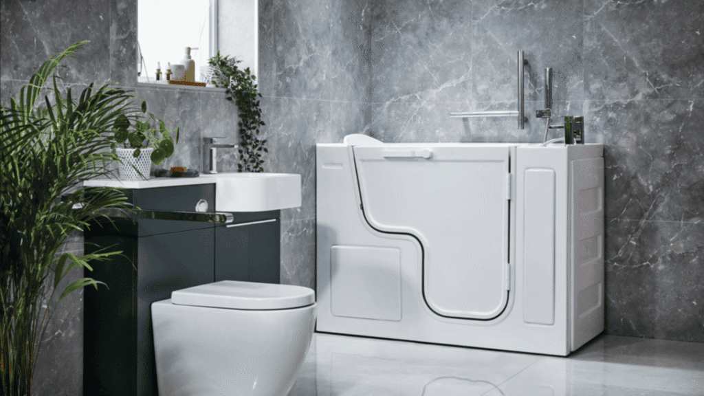 A high-resolution hero image of a contemporary accessible bathroom featuring a premium white walk-in bath with a uniquely shaped, low-threshold door and integrated chrome grab rails. The room is finished with luxury grey marble-effect wall tiles and includes a modern white toilet and a compact washbasin set into a dark grey vanity unit. Natural light from a window and lush greenery from a potted palm and hanging plants create a sophisticated, spa-like environment that prioritises both safety and style.