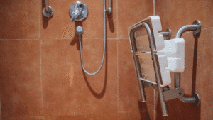 A close-up view of a white and chrome fold-down accessible shower seat, shown folded up against a textured terracotta-tiled shower wall. Chrome shower fixtures are visible in the background. Practical and safe.